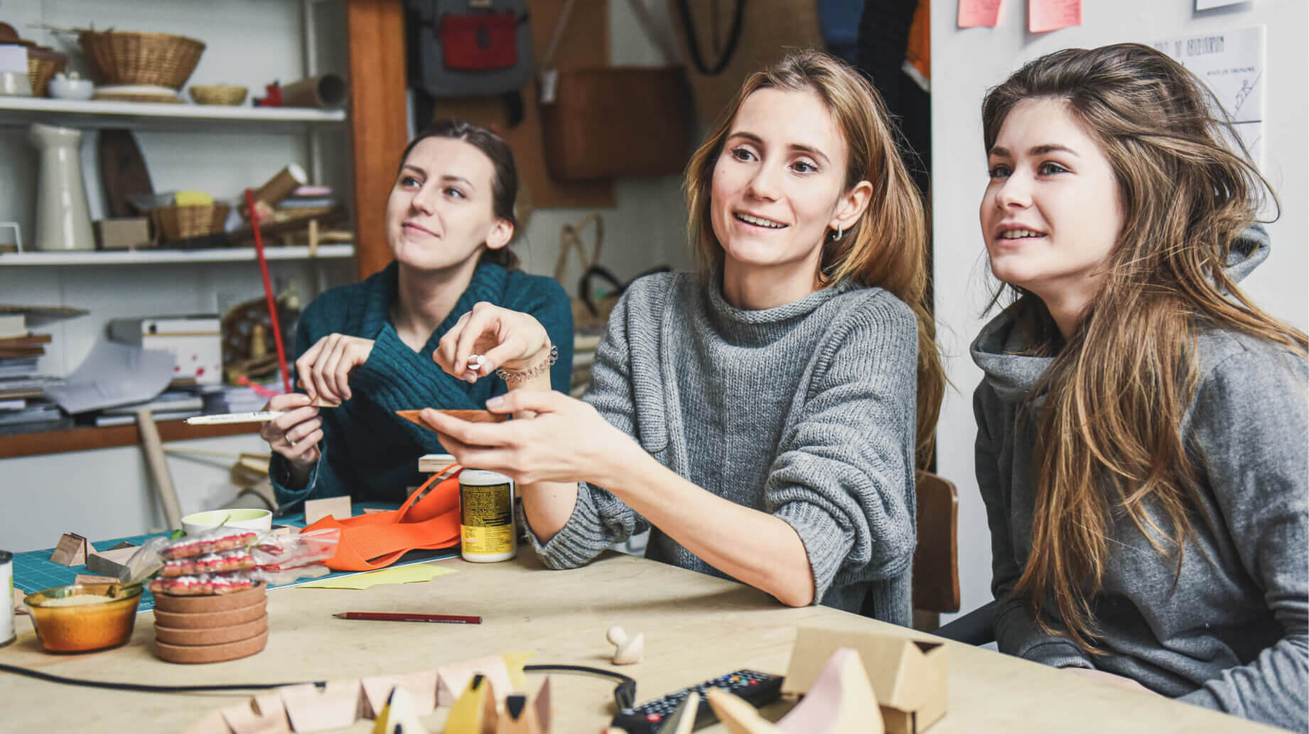 Trzy studentki podczas zajęć. Siedzą przy drewnianym stole na którym leżą fragmenty drewnianych zabawek i akcesoria do modelowania. W tle drewniane półki z plecionymi koszami, ceramiką, obiektami ze skóry.