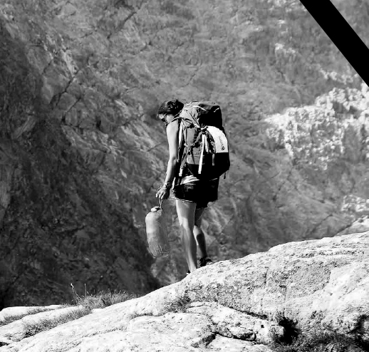 Czarno-biała fotografia Małgosi Kęckiej stojącej na skale, z plecakiem na plecach i śpiworem w dłoni. W tle ściana skalna.